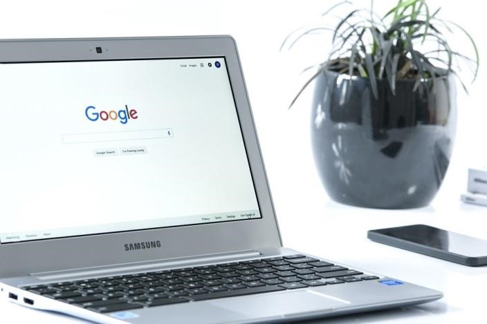 A silver laptop on a white desk next to an indoor plant and smartphone, displaying Google’s search engine page