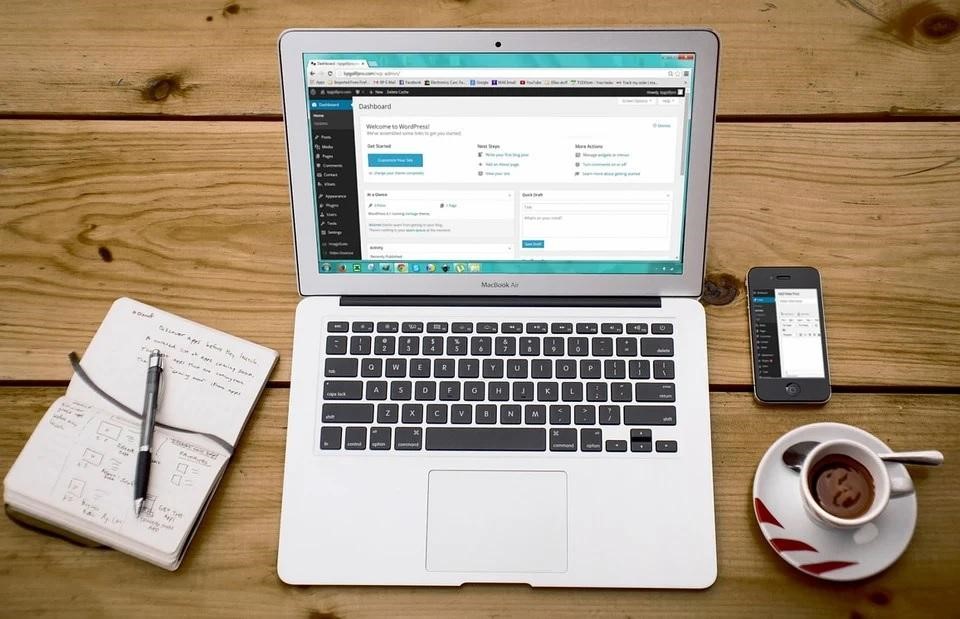 A silver laptop on a wooden desk, next to a notebook, smartphone, and coffee mug